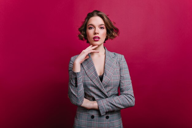 Interested businesswoman with trendy makeup posing on claret wall. Indoor photo of serious young lady in tweed jacket standing in confident pose.