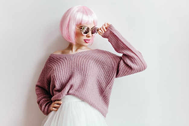 Interested caucasian girl in purple sweater and white skirt standing near wall. adorable young woman in pink periwig and sunglasses posing on light wall.