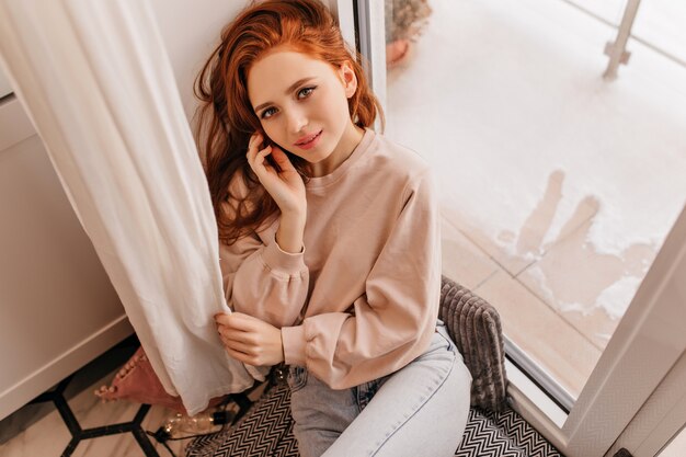 Interested caucasian girl sitting on window sill. Relaxed ginger woman chilling at home.