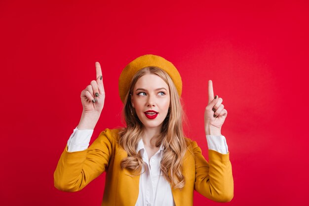 Interested caucasian lady in yellow beret pointing up with fingers. Cute french girl gesturing on red wall.