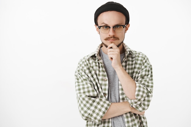 interested creative bearded young man in black beanie and glasses holding hand on chin and gazing with serious thoughtful expression thinking listening curious plan