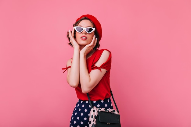 Interested cute woman in white sunglasses looking up Studio portrait of relaxed caucasian girl in red french beret