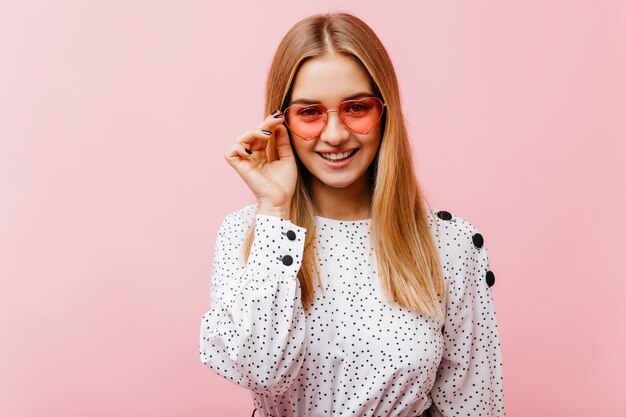 Interested european woman in sunglasses standing on pink. Fascinating woman in white blouse having fun