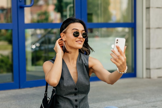 灰色のベストと濃いサングラスに興味のある魅力的な女性がスマートフォンで自分撮りを作っています