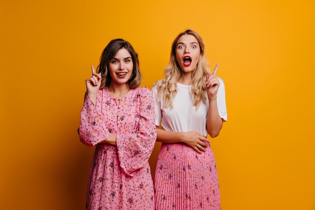 Interested ladies in trendy clothes posing on bright wall. Portrait of curious female friends standing in front of yellow wall.