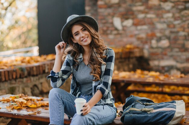 Interested lady with elegant black manicure drinks coffee on wooden bench with golden leaves on background