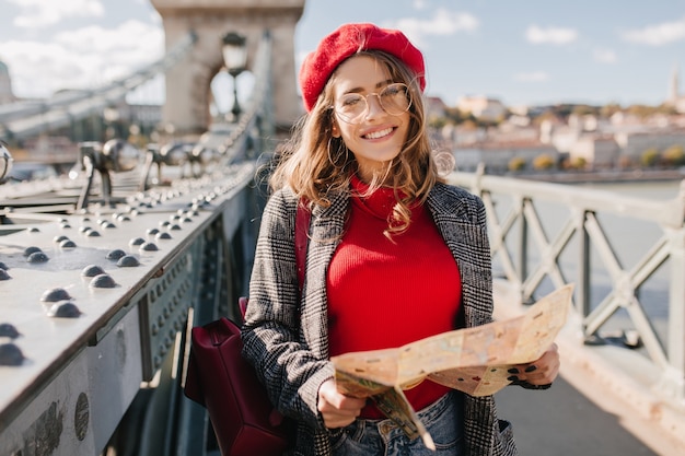 Foto gratuita donna bianca interessata in maglione rosso e berretto che trascorre del tempo all'aperto, esplorando la città con la mappa