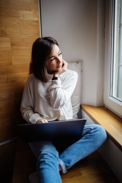 Interested woman is working on laptop while sitting on wide windowhill in daily time