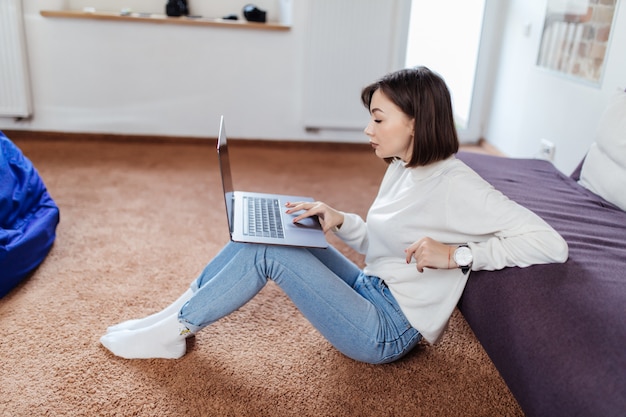 興味がある女性は黒いソファの近くの床に座っているラップトップコンピューターで動作します