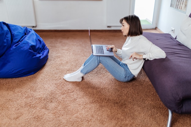 Interested woman works on laptop computer sitting on the floor near black sofa