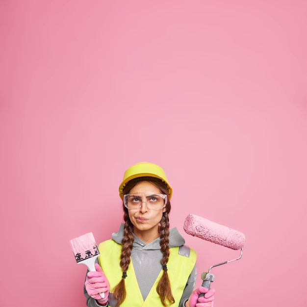 Interior renovation and home improvement. Serious displeased woman dressed in building clothes holds repair tools focused above ready for painting walls isolated over pink wall copy space