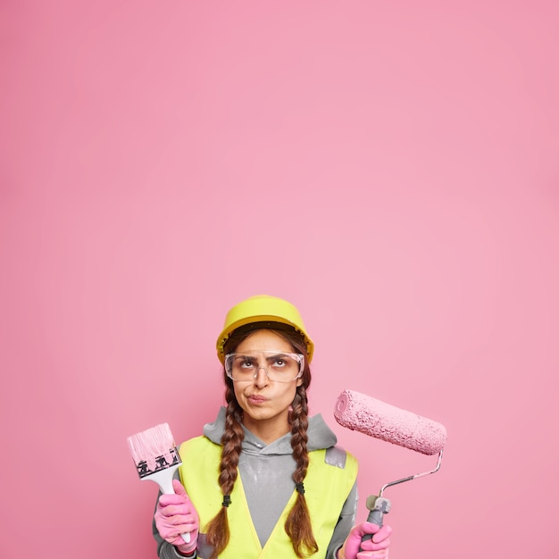 インテリアのリノベーションと家のリフォーム。建物の服を着た深刻な不機嫌な女性は、ピンクの壁のコピースペースに隔離された壁をペイントする準備ができている上に焦点を合わせた修理ツールを保持