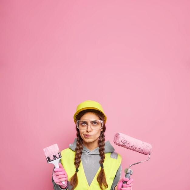 Interior renovation and home improvement. Serious displeased woman dressed in building clothes holds repair tools focused above ready for painting walls isolated over pink wall copy space