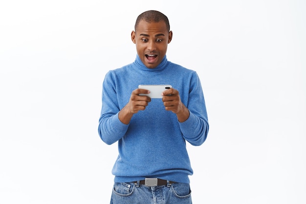 Internet online lifestyle and people concept Portrait of excited happy young african american adult guy playing racing game watching movie or video on smartphone hold mobile phone both hands