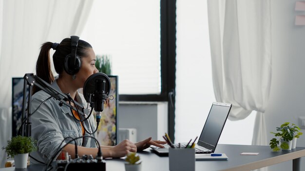 Internet radio show host talking on microphone while checking subject list on laptop. Popular social media content creator speaking to audience while sitting in modern studio.