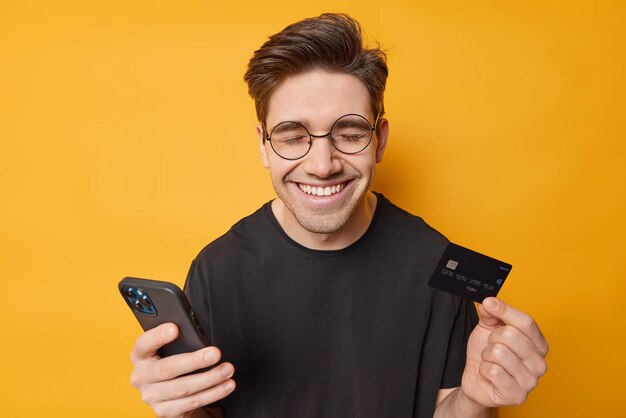 インターネットショッピングのコンセプト幸せな黒髪の若い男は携帯電話を使用し、クレジットカードは黄色の背景の上に分離された黒いTシャツに身を包んだアプリケーションを使用してオンライン購入を行います大セール