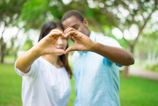 Interracial couple joining fingers in shape of heart while having date in summer park.