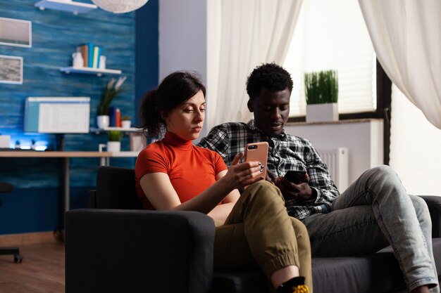 가제트와 함께 편안한 소파에 앉아 인종 커플입니다. 기술로 스마트폰을 사용하는 백인 여성과 아프리카계 미국인 남성. 전화 장치를 들고 있는 혼혈 파트너.