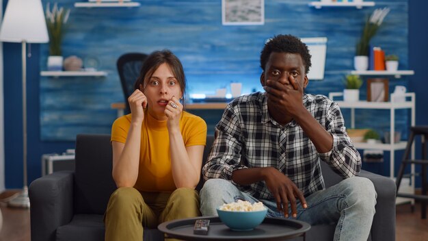 Межрасовая пара смотрит драматический фильм по телевидению в гостиной. молодые многоэтнические партнеры в шоке, глядя в камеру на диване. смешанная раса муж и жена дома