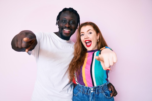 Interracial couple wearing casual clothes pointing to you and the camera with fingers, smiling positive and cheerful