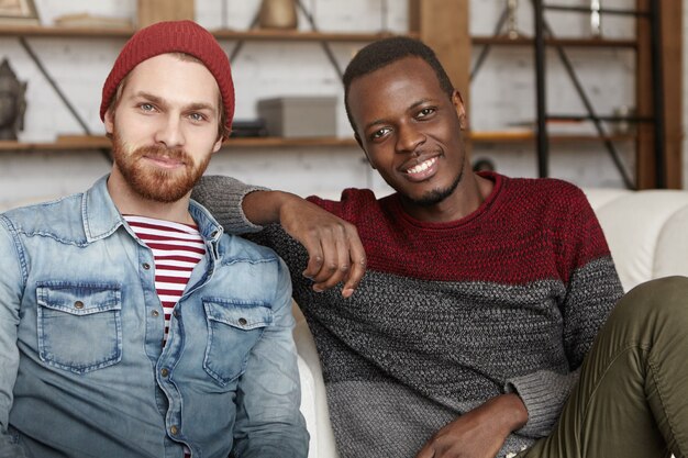 Interracial friendship concept. Happy African American male in casual sweater resting elbow on his best friend's shoulder while sitting on white couch at coffee shop, talking and having fun together