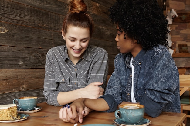 Foto gratuita coppia lesbica interrazziale guardando in basso con un sorriso timido, tenendosi per mano durante il pranzo al ristorante