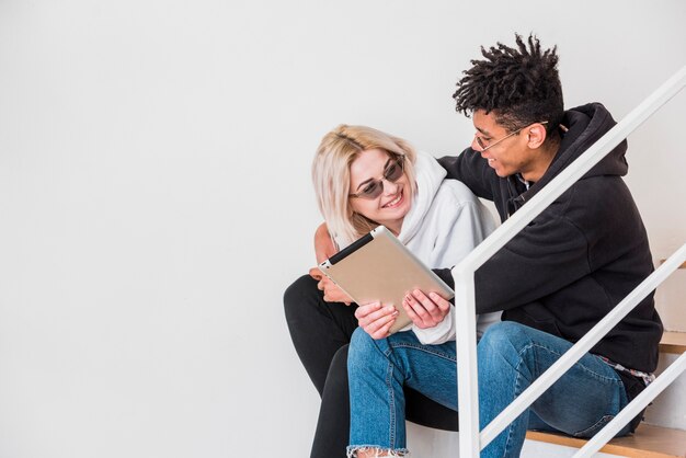 An interracial young couple sitting on staircase using digital tablet against white wall