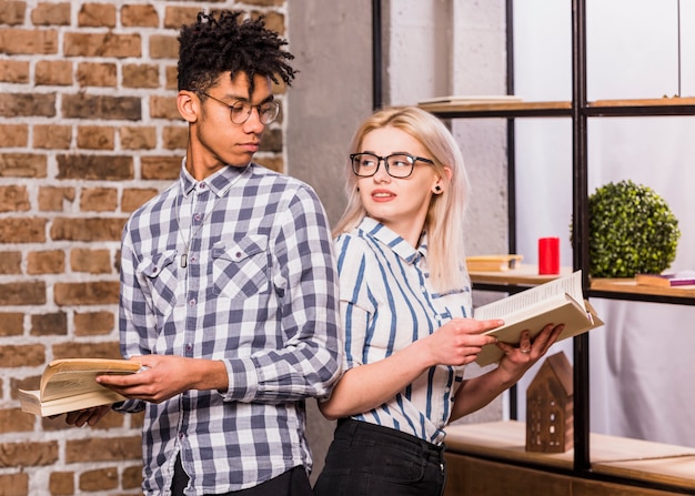 Free photo an interracial young couple standing back to back holding books in hand looking at each other