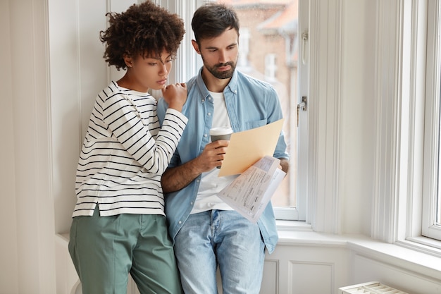 Interracial young woman and man make accounting of their expenses for year, concentrated on paper work