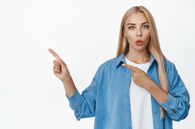 Intrigued blond girl pointing fingers left, asking question about company product, showing something interesting on copy space on white