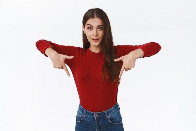 Заинтригованная любопытная красивая брюнетка в красном свитере заинтересована в какой-то промо, задает вопрос и указывает вниз, поднимает одну бровь, указывая на нижнюю рекламу на белом фоне