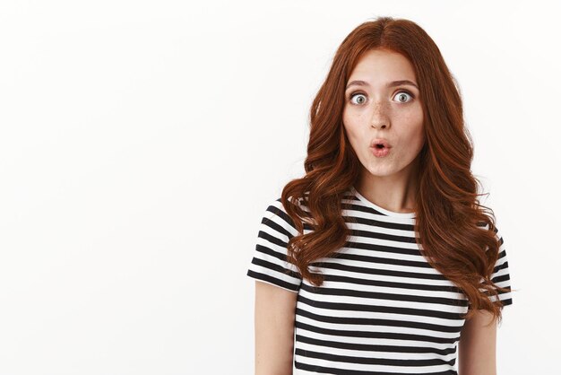 Intrigued cute silly redhead woman in striped tshirt whistling from excitement and interest pouting stare astonished like gossiping hear entertaining news standing white background
