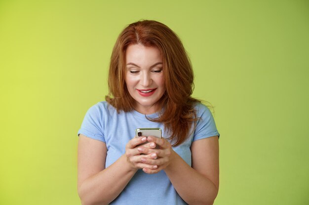 興味をそそられる興奮した魅力的な赤毛中年女性のショッピングオンラインスマートフォンホールド携帯電話見て喜んで楽しませてくれたガジェット画面笑顔喜んで遊んでいるゲームが完璧なアプリを見つけました
