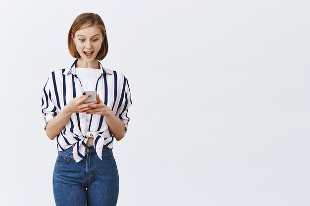 Заинтригованная и взволнованная молодая женщина проверяет сообщения или банковский счет по телефону, изумленно глядя на смартфон