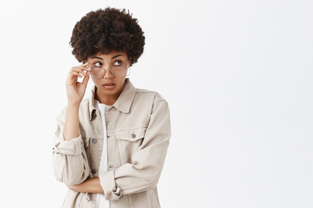 Intrigued good-looking african american, looking from under glasses, touching rim of eyewear and staring right with clueless expression over gray wall