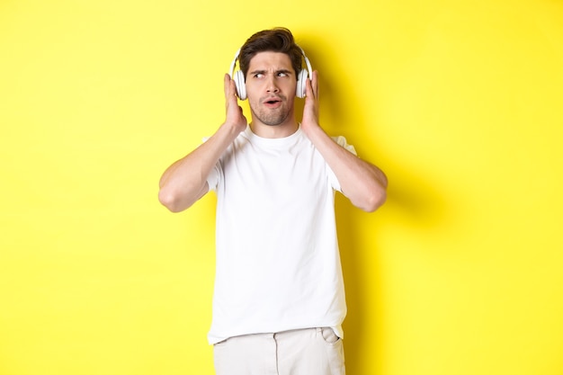 Intrigued guy enjoying tunes in headphones, listening closely to music in earphones, standing over yellow background.