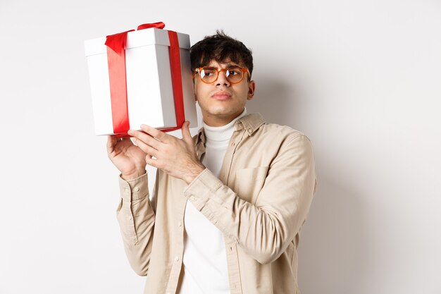 Intrigued guy listening whats inside gift box, trying guess present, standing focused on white background.