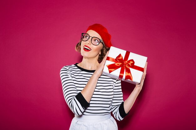 Intrigued happy ginger woman in eyeglasses holding gift box near the ear and looking up over pink