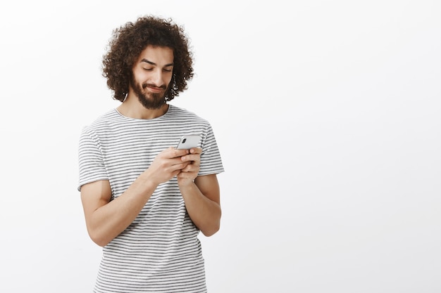 intrigued joyful hot hispanic guy with beard and afro haircut, smiling at screen of smartphone