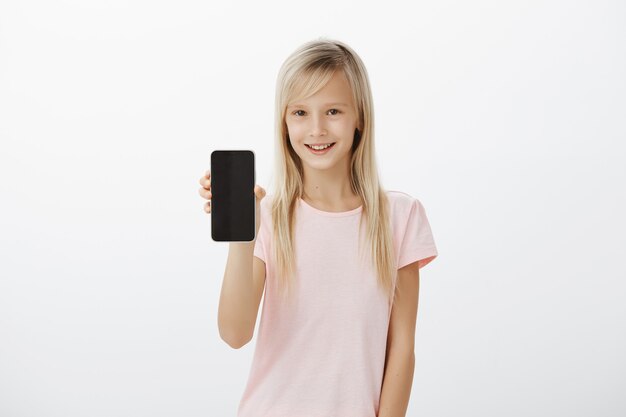 intrigued neat blond girl in pink t-shirt, smiling curiously and showing black smartphone