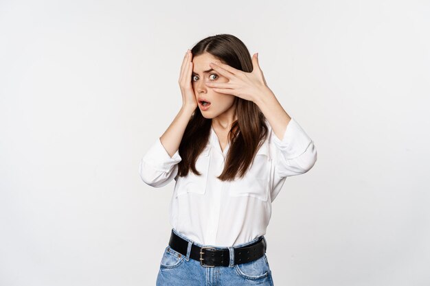 Intrigued shocked young woman peeking through fingers at smth interesting standing over white backgr...