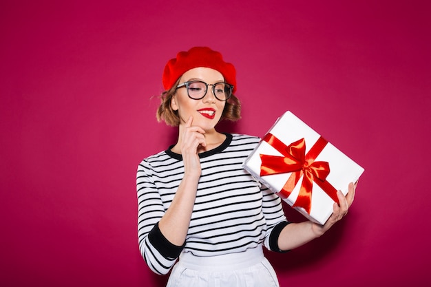 Заинтригованная улыбающаяся рыжая женщина в очках держит и смотрит на подарочную коробку на розовом