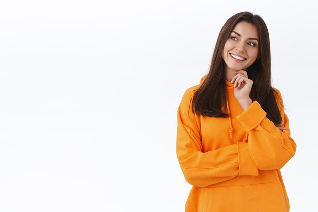 Intrigued and thoughtful caucasian girl in orange hoodie touch chin tilt head and looking left curious smiling interested contemplate cool thing making choice standing white background ponder