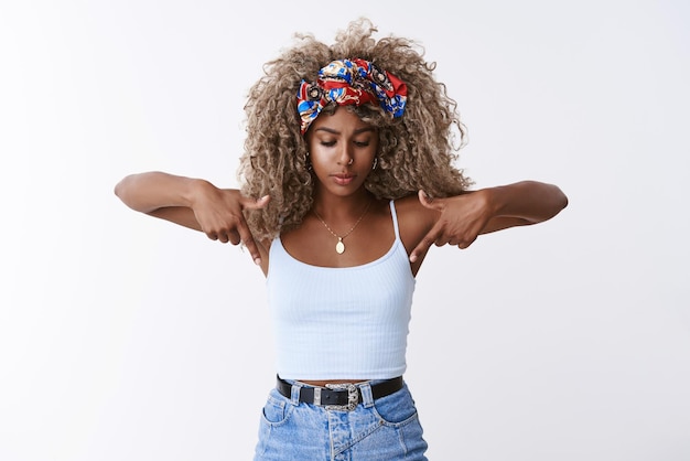 Intrigued and thoughtful seriouslooking blond curlyhaired hipster woman with stylish headband bend pointing and looking down concentrated standing white background staring something