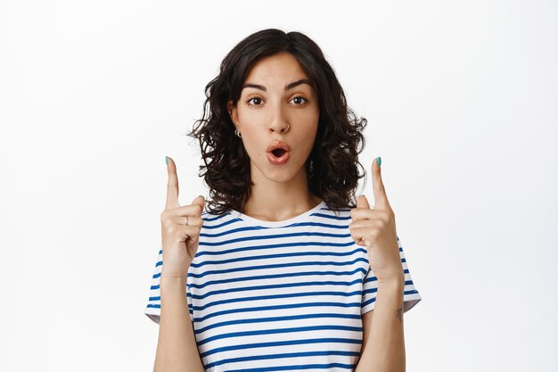 Intrigued young brunette woman, jewish girl pointing fingers up and say wow, look impressed and excited with special discounts, offer in shop, standing  on white