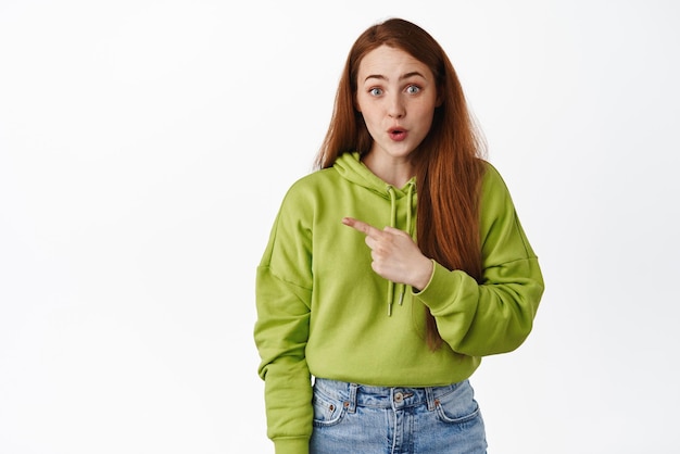 Intrigued young redhead girl pointing finger left look excited with advertisement showing promo offer standing against white background