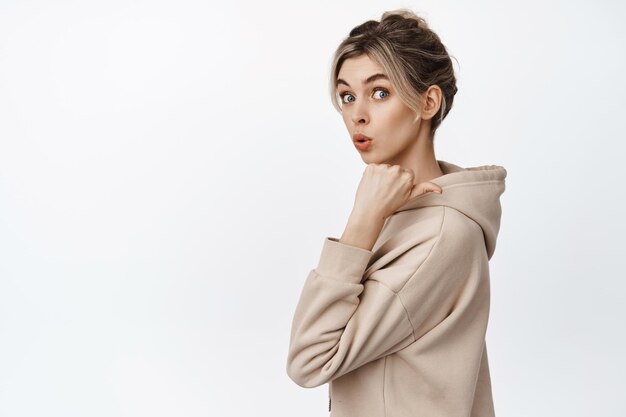 Intrigued young woman pointing finger right looking surprised and curious showing advertisement behind her standing over white background