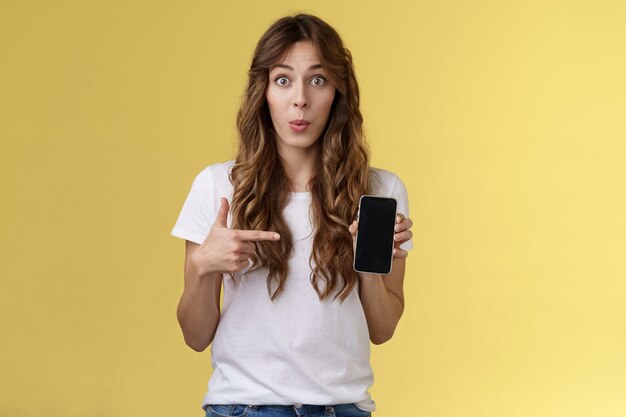 Intriguing app check out. Enthusiastic surprised attractive girlfriend gossiping friend new boyfriend showing curious photo smartphone hold mobile phone pointing telephone screen yellow background