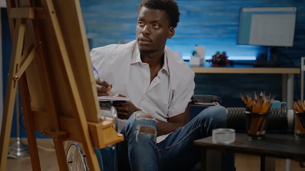 Invalid artist of african american ethnicity working on sketch for modern drawing in studio room at home. Black young person sitting in wheelchair while designing masterpiece for art project