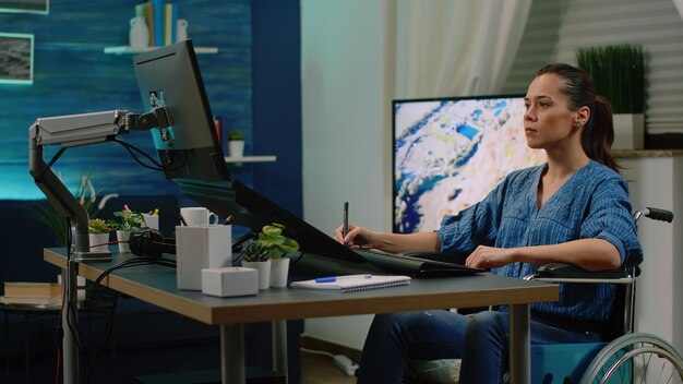 スタジオのコンピューターでグラフィックタブレットとスタイラスを操作している無効な写真家。タッチスクリーンで写真をレタッチし、車椅子に座っているハンディキャップを持つ写真家。